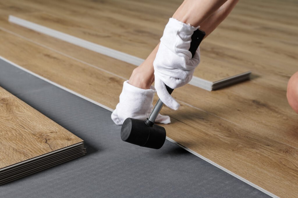 Close view of hands aligning SPC vinyl plank on acoustic underlayment with a rubber mallet during flooring installation
